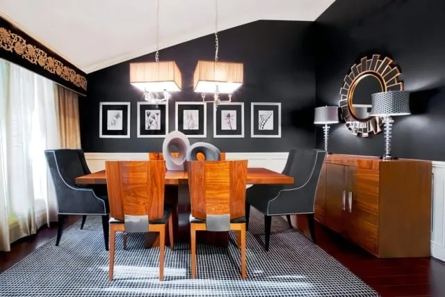 Modern dining room with a wood table, six chairs, and a black-and-white rug. A black accent wall showcases framed artwork and a round mirror perched above a sideboard. Pendant lights float overhead, while large, curtained windows flood the space with light—a testament to new home interior design in Long Island.