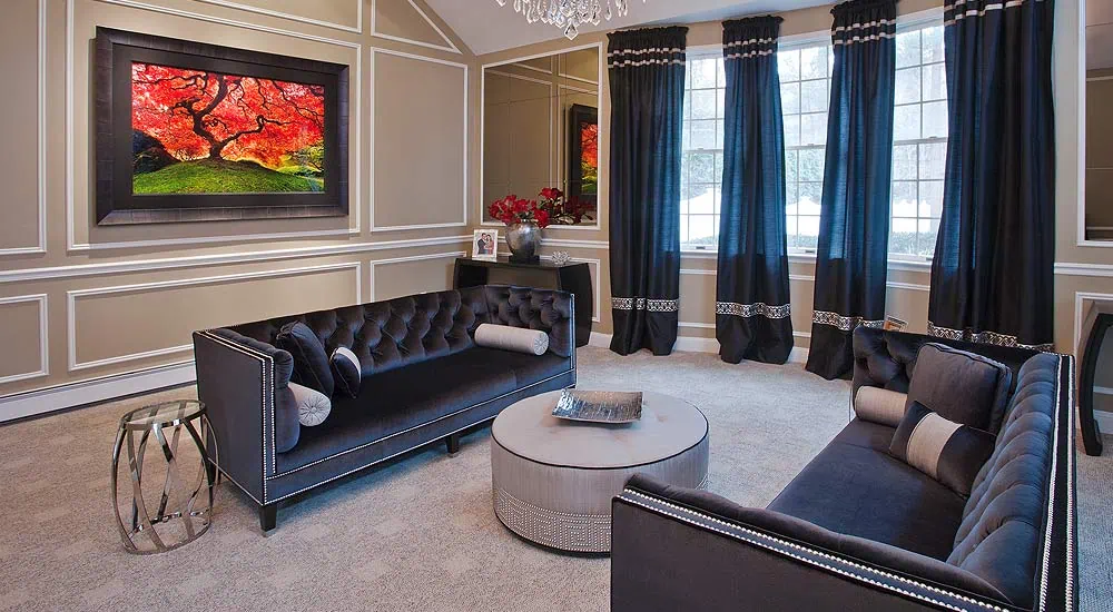 A sophisticated living room in Long Island, NY, displays dark blue velvet sofas and a round gray coffee table. Interior designers skillfully incorporate elegant wall moldings and vibrant artwork with red and green hues. A large window with blue curtains bathes the space in natural light.