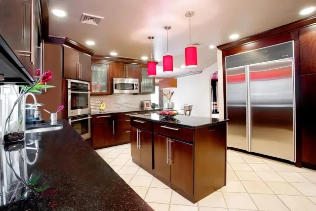 Modern NY kitchen with dark wooden cabinets, stainless steel appliances, and a large silver fridge. An island with a black countertop and red pendant lights takes center stage. A tile floor and decorative vase add elegance, embodying the expertise of Long Island interior designers.