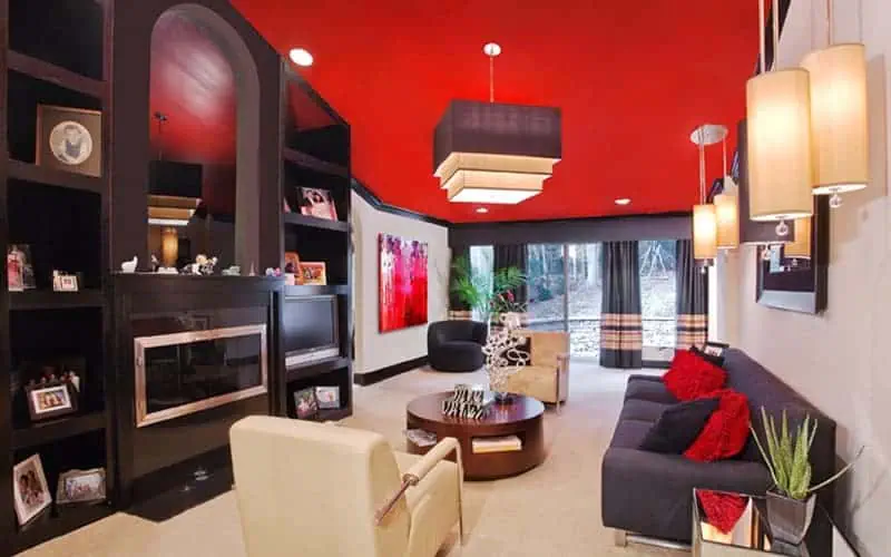 This modern living room, crafted by top interior designers from Long Island, NY, boasts a bold red ceiling, black and white furniture, and a wall-mounted fireplace. Contemporary decor with abstract artwork is illuminated by hanging lamps, while large windows offer garden views.