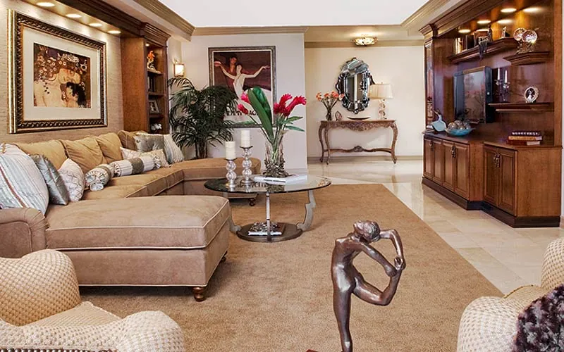 Elegant living room with a beige sectional sofa, colorful pillows, and a glass coffee table adorned with red flowers. Art and sculptures decorate the space alongside wooden cabinets and a large wall mirror. Neutral tones create a warm atmosphere in this new home interior design on Long Island.