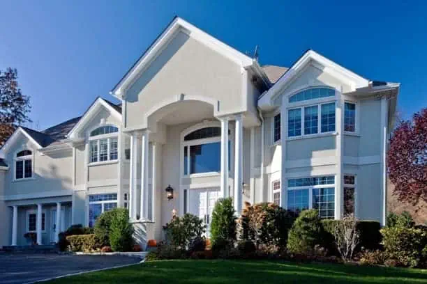 Large white house with tall columns and multiple windows under a clear blue sky. Located in Long Island, NY, it's graced by a front yard with a well-maintained lawn and autumnal orange accents. Sunlight casts shadows, highlighting the building's architectural details and sophisticated interior design.