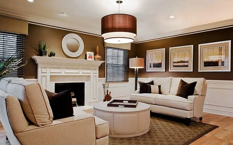 A stylish living room in a new home on Long Island features beige sofas with dark cushions, a round coffee table, and a white fireplace. A large brown pendant light hangs above. Artwork and a round mirror adorn the brown walls, while windows with black blinds allow natural light to illuminate the space.