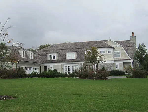 This large two-story house in NY features gray siding and a shingled roof, surrounded by trees and a lush green lawn. The overcast sky suggests a cloudy day, enhancing its classic architecture with dormer windows and a prominent chimney, ideal for those seeking new home interior design on Long Island.