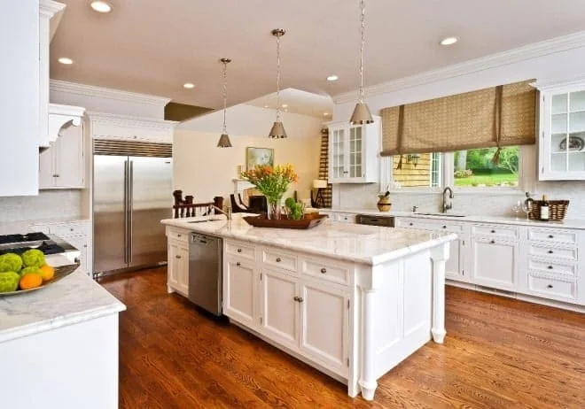 A spacious kitchen with white cabinetry, marble countertops, and a large island designed by interior designers from Long Island. The island holds a tray with a vase of flowers. Stainless steel appliances and pendant lights add a modern touch, while wooden floors complete the warm ambiance.