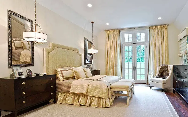 A luxurious bedroom in a new home interior design on Long Island features a large bed with an upholstered headboard, flanked by two nightstands with lamps. Elegant curtains frame French doors leading outside, while a plush chair and dresser complete the tasteful decor envisioned by top interior designers from NY.