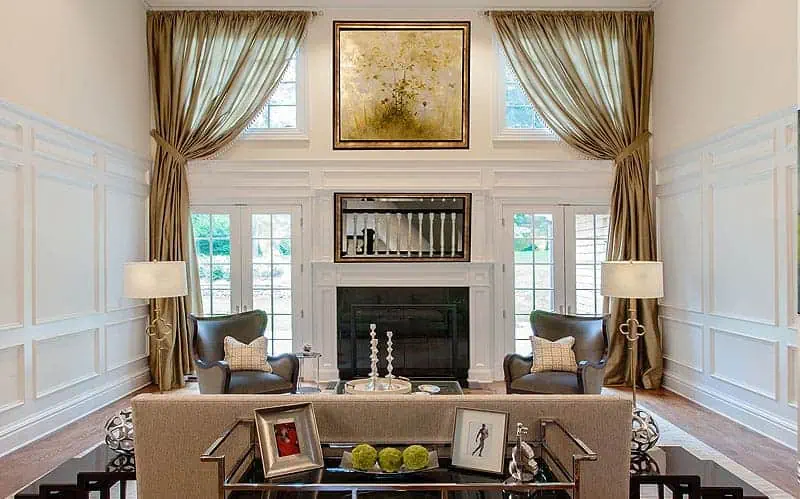 Experience the epitome of luxurious new home interior design on Long Island in this stunning living room. High ceilings, large windows with elegant drapes, and a fireplace crowned by art create a sophisticated ambiance. Classic paneling encases two armchairs and a chic glass coffee table.