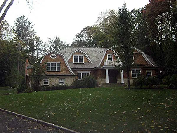 A large two-story house with a shingled roof and gabled roofline sits amidst a landscaped lawn, perfect for those seeking new home interior design in Long Island. With multiple windows and a front porch, it’s surrounded by trees and bushes under overcast skies.