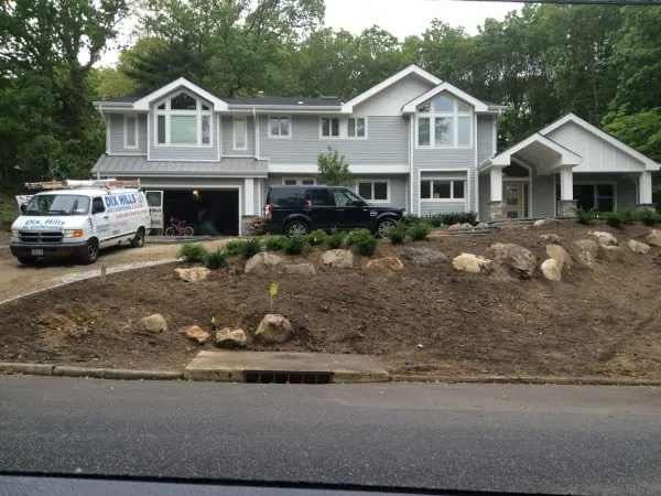 A two-story gray house with large windows nestles among the trees, reflecting the charm of Long Island living. Outside, a white van with "Dew Mills" and a black SUV sit in the driveway. The front yard showcases newly planted shrubs and rocks on a sloped, barren patch of land.