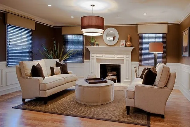 A cozy living room in NY with brown walls and wooden floors boasts two beige sofas facing each other, a round coffee table nestled between them, and a white fireplace. Embracing new home interior design Long Island style, the mantel is adorned with a round mirror and decor items. Two lamps and large windows illuminate the space.