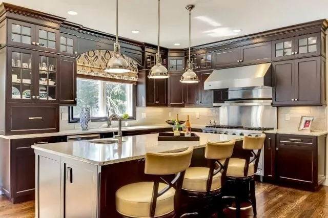 A modern kitchen in a new home interior design on Long Island, NY features wooden cabinets, stainless steel appliances, and a central island with three cushioned chairs. Pendant lights hang above the island, and a large window bathes the space in natural light. A bottle of wine and a vase adorn the counter.