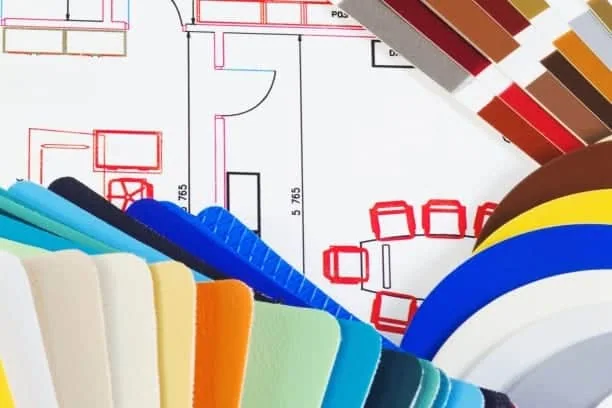 A collection of multicolored paint swatches is fanned out over an architectural floor plan. Perfect for inspiring interior designers in Long Island, the plan features red and black lines depicting furniture and measurements. The swatches include a variety of colors and textures.