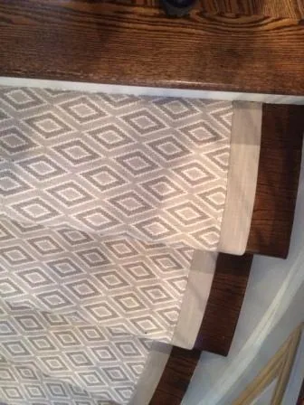 Close-up of a carpet runner on wooden stairs, ideal for those seeking new home interior design in Long Island, NY. The runner showcases a geometric diamond pattern in beige and white, secured along dark brown steps, offering a stylish touch admired by interior designers in Long Island.