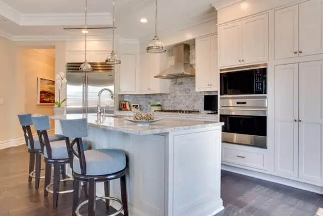This modern kitchen in a new home interior design on Long Island features white cabinetry, a large island with marble countertops, and three blue upholstered bar stools. Stainless steel appliances, including a fridge and built-in ovens, seamlessly integrate into the design as pendant lights illuminate the space.