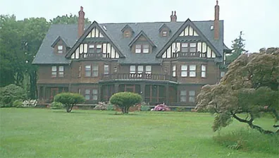 A large, historic Tudor-style mansion with a dark roof and exposed wooden beams on the upper floors graces the lush greenery of Long Island. The foreground showcases a well-maintained lawn, hinting at the exquisite new home interior design tailored by skilled interior designers.
