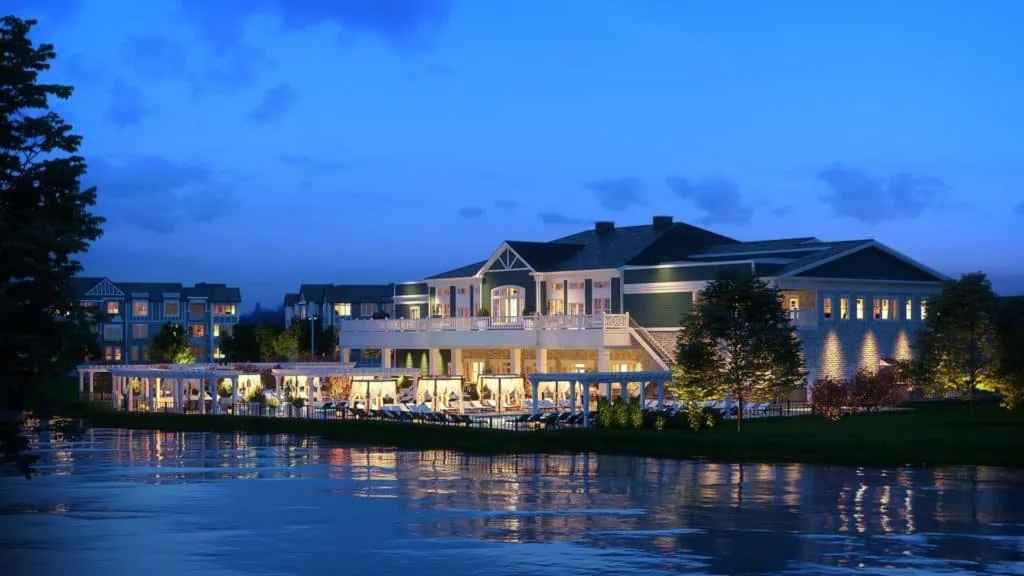 A luxurious, well-lit clubhouse by the water at dusk boasts large windows and a spacious balcony, designed by renowned interior designers from Long Island. Trees and landscaped greenery surround the serene area, reflecting in the calm water under the deep blue NY sky.