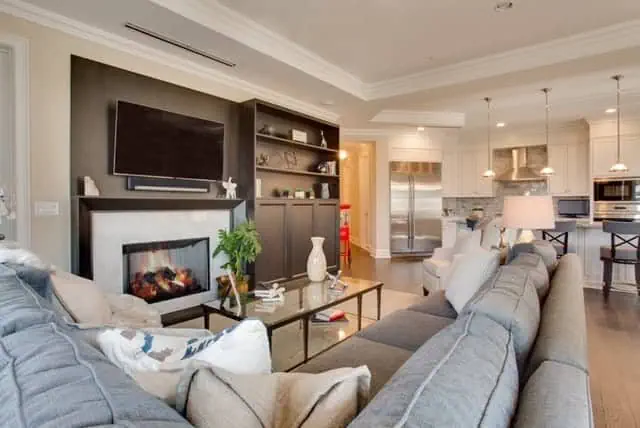 Spacious living room featuring a gray sectional sofa, coffee table, and fireplace with a wall-mounted TV above. This beautiful new home interior design on Long Island also boasts an open kitchen with bar stools, stainless steel appliances, and pendant lights, all in calming neutral tones.