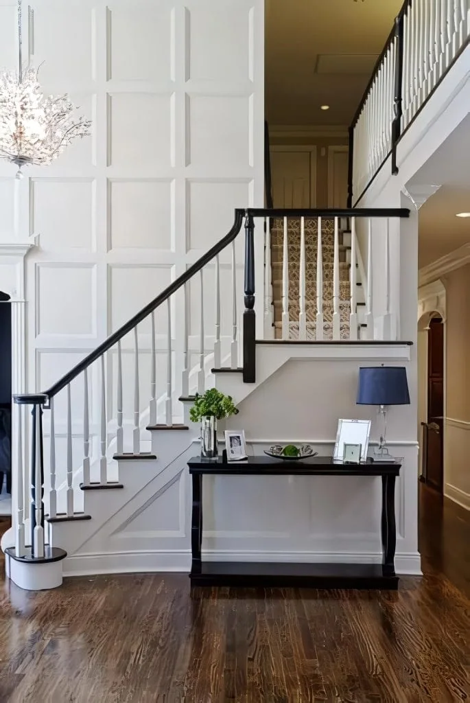 Spacious foyer with dark wood floors and white walls, featuring a staircase with a black handrail. A table adorned by skilled interior designers from Long Island showcases framed photos, a lamp, and flowers beneath the stairs. A chandelier gracefully hangs from the ceiling.