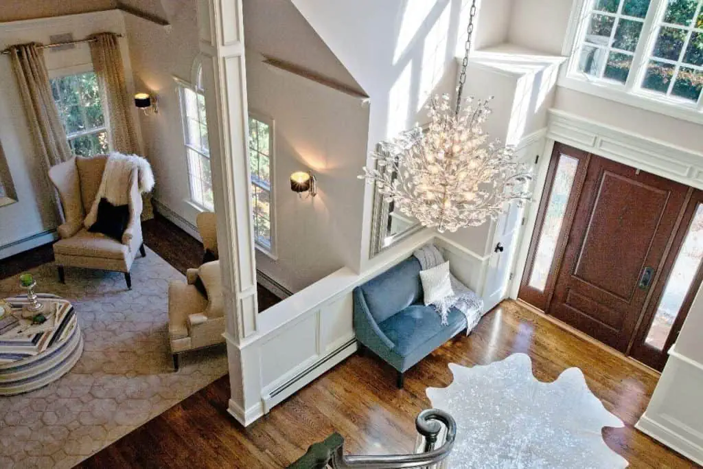Overhead view of a stylish living room in a new home interior design in Long Island, NY. Featuring a chandelier, wooden floors, and a large rug. A sitting area with two chairs and a table is on the left, while a plush bench sits near the wooden front door on the right. Natural light streams through tall windows.