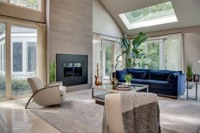 Modern living room with a high ceiling and large window designed by top interior designers in Long Island. Features a blue sofa, armchairs, and a glass coffee table. A fireplace graces the wall, while plants enhance the space's charm. Skylights and glass doors fill this new home interior design with natural light.