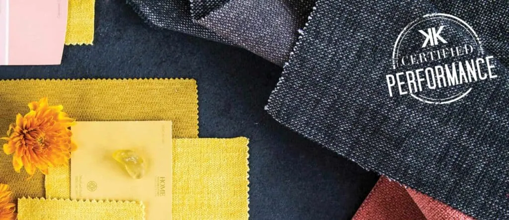 Swatches of fabric in various colors, including yellow, pink, and black, are arranged on a dark surface alongside two yellow flowers. A stamp reading "Certified Performance" is visible on one fabric—a perfect palette for interior designers crafting elegant new home designs in Long Island, NY.