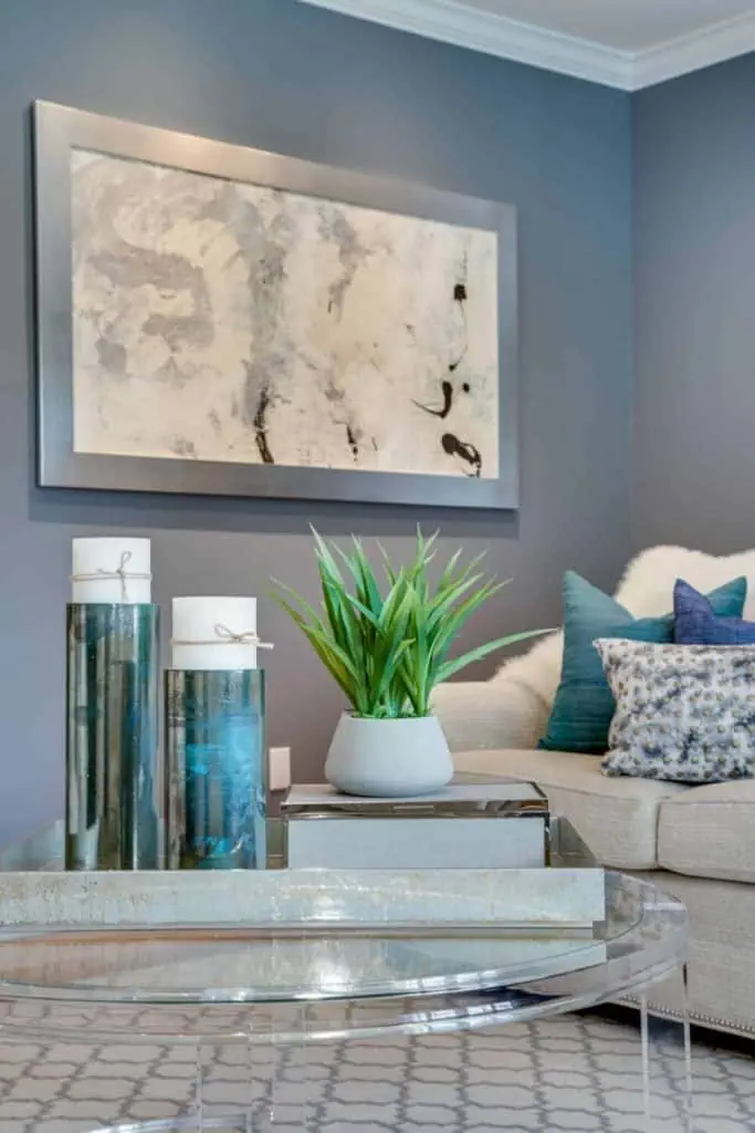 A contemporary living room in Long Island, NY, boasts blue walls and a large abstract painting. A glass coffee table displays decorative vases and a potted plant, while a cream-colored sofa with patterned cushions invites comfort—a perfect example of refined new home interior design.