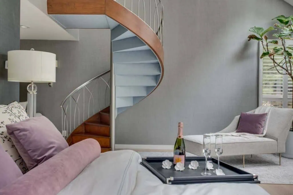 A cozy NY bedroom with a modern spiral staircase features a large bed with purple pillows and a tray holding champagne. A plush chair near the window with a leafy plant complements the soft gray walls, showcasing the elegant touch of interior designers from Long Island.