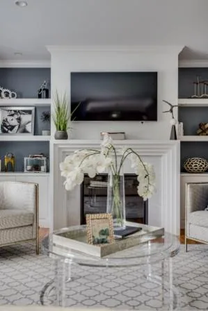Living room in a new home on Long Island, showcasing a wall-mounted TV above a white fireplace. Shelves on both sides display decor items. A glass coffee table with a vase of white flowers and a framed picture sits between patterned armchairs atop a geometric-patterned rug.