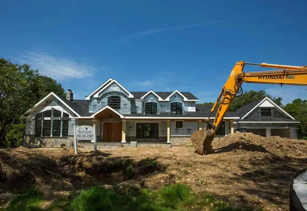 A large, partially constructed house with a gray stone facade and a steep roof stands behind a mound of dirt in Long Island, NY. An excavator is parked in front, signaling progress on this promising new home. A signboard with contact information hints at the stylish interior design awaiting within. The sky is clear and blue.