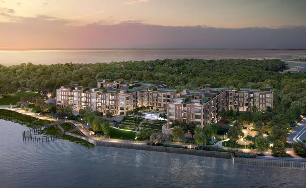 Aerial view of a large, modern apartment complex surrounded by lush greenery along a NY waterfront. The buildings are illuminated, and the scene is set during sunset with a serene body of water in the foreground, showcasing the sophisticated touch of interior designers from Long Island.