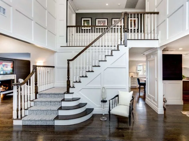 A spacious, modern home interior crafted by renowned interior designers from Long Island features a grand staircase with white paneling and dark wood accents. A curved step leads to the wooden floor, complemented by a white chair and lamp. Artwork and a fireplace grace the adjoining rooms.