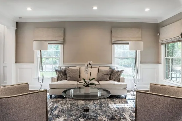 A sophisticated living room designed by interior designers from Long Island features a beige sofa adorned with gray pillows, flanked by two tall lamps. A glass coffee table sits on a patterned rug, and windows with beige blinds allow natural light to filter into this elegant new home.