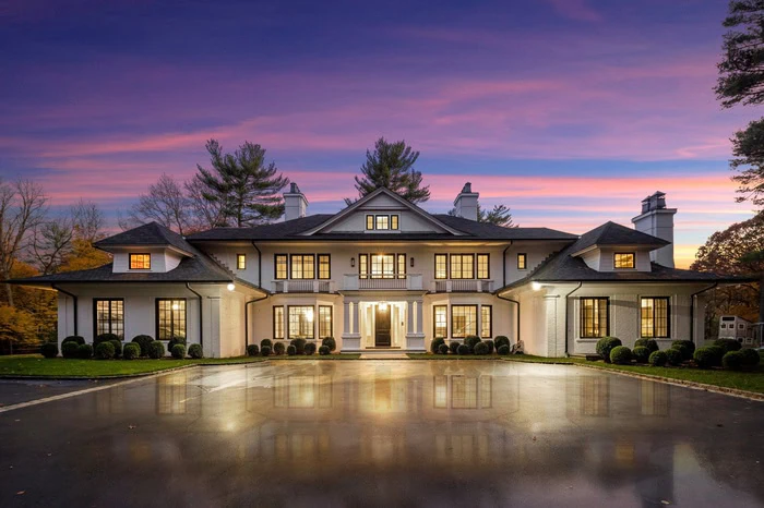 A large, elegant two-story house with numerous windows reflects on a wet driveway, showcasing the charm of new home interior design Long Island is renowned for. The sky is a vibrant mix of purple and pink hues at sunset, while trees surround the property.