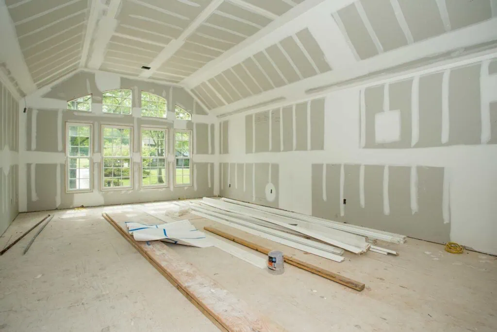 A bright, unfinished room with drywall installation in progress. Sunlight streams through large arched windows, illuminating scattered wooden planks and a paint can on the floor. The vaulted ceiling adds height to the space, perfect for interior designers Long Island to transform into a stunning New York retreat.