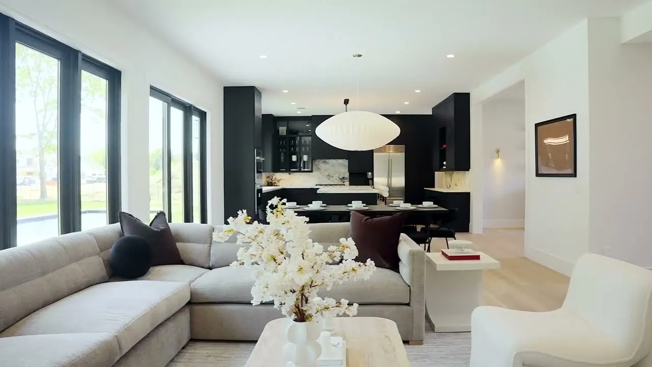 This modern living room showcases new home interior design with a light gray sectional sofa, white flowers on a wooden coffee table, and artistic wall decor. The open-concept space flows into a sleek black kitchen. Designed by talented interior designers from Long Island, large windows illuminate the room with natural light.