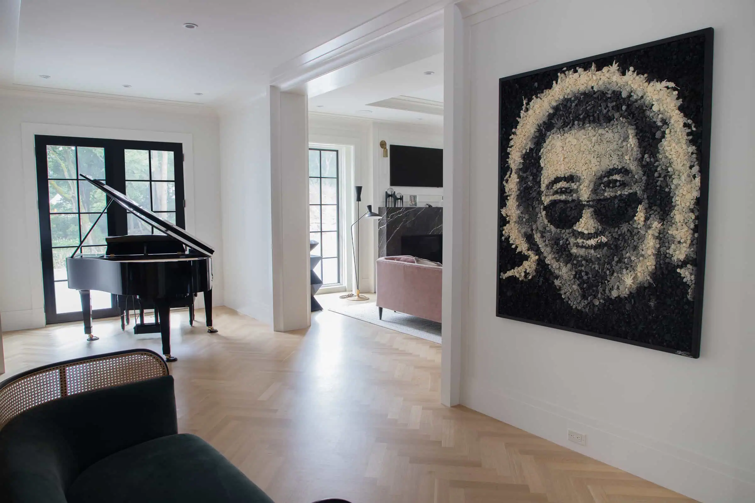 Spacious living room in a NY home, featuring a grand piano and large windows. A striking portrait of a bearded face wearing sunglasses hangs prominently on the wall. Elegantly designed with light wood flooring and modern furniture, this interior showcases Long Island's finest design inspiration.