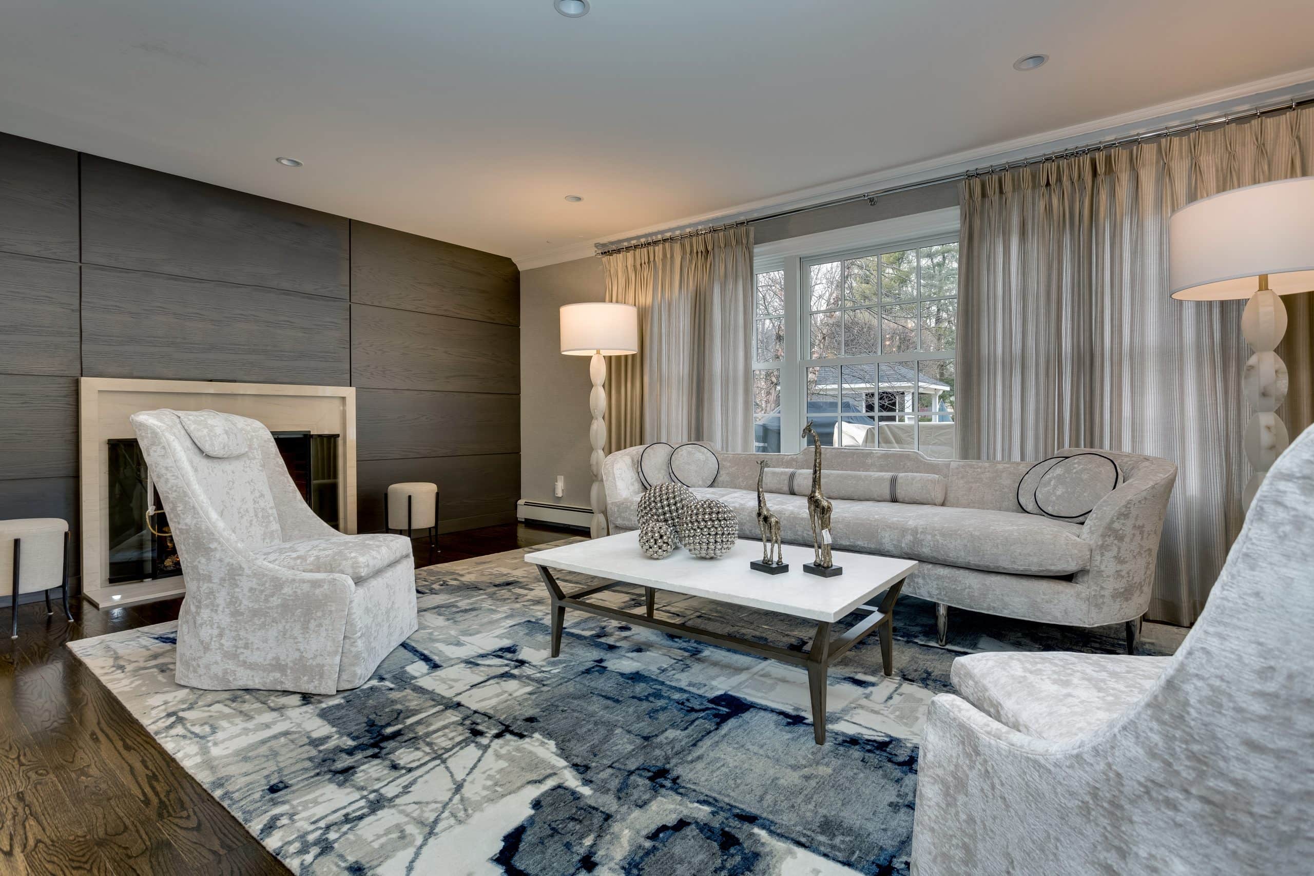 Modern living room with neutral-toned furniture, a patterned blue and white rug, two tall lamps, a coffee table with decorative items, large window with curtains, and a sleek fireplace.