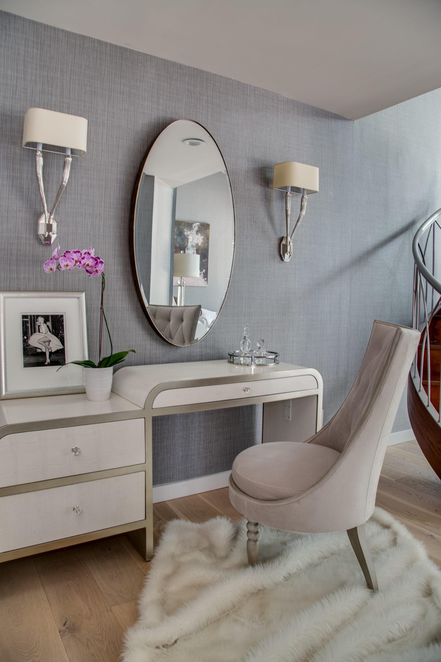 A modern vanity area with a white desk, oval mirror, upholstered chair, framed photo, potted orchid, and glass decanters. Two wall sconces flank the mirror. The floor has a white rug.