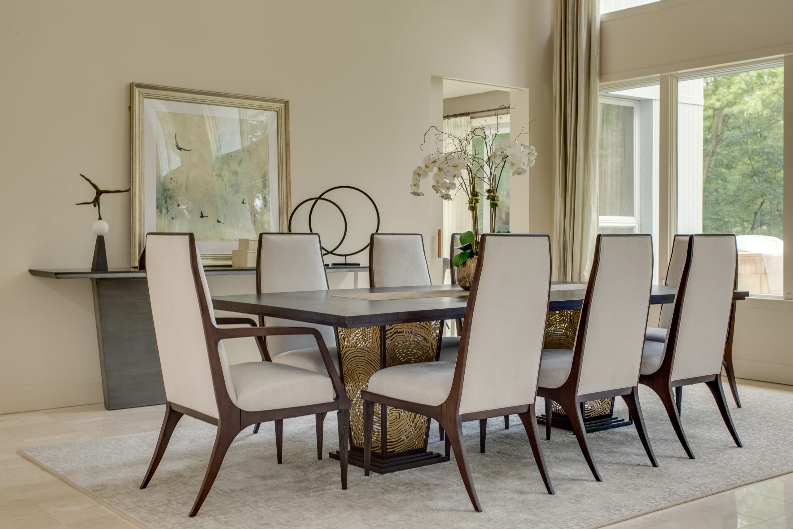 A modern dining room with a rectangular table, eight upholstered chairs with dark wooden frames, a light area rug, decorative art on the wall, and large windows letting in natural light.