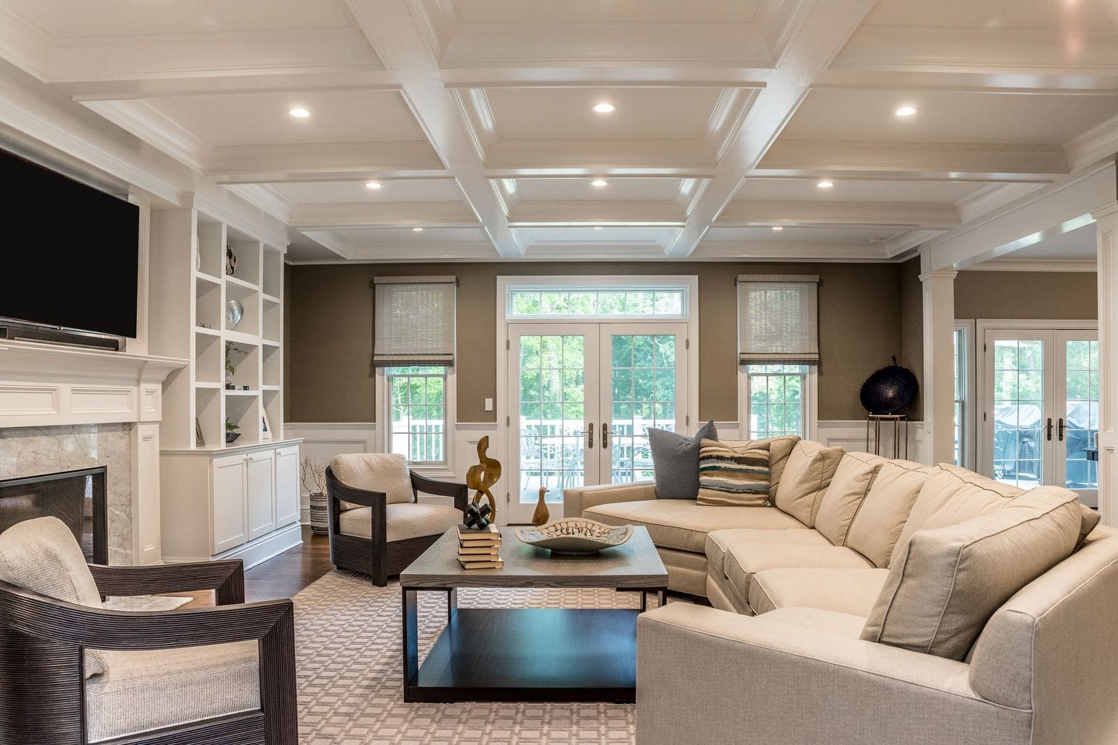 A spacious, modern living room with a large beige sectional sofa, two armchairs, a coffee table, built-in shelves, a fireplace, and large windows letting in natural light.