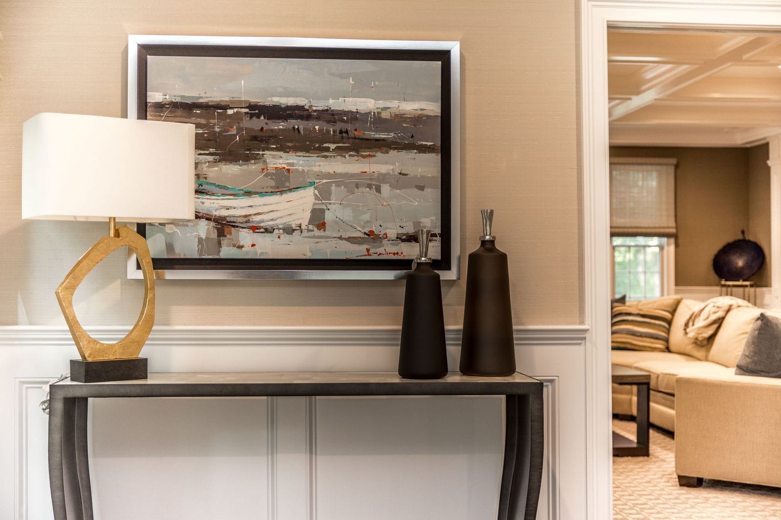 A modern console table with a sculptural lamp, two tall black vases, and a framed abstract painting of boats hangs on the wall; in the background, a cozy living room with a sofa and patterned carpet is visible.