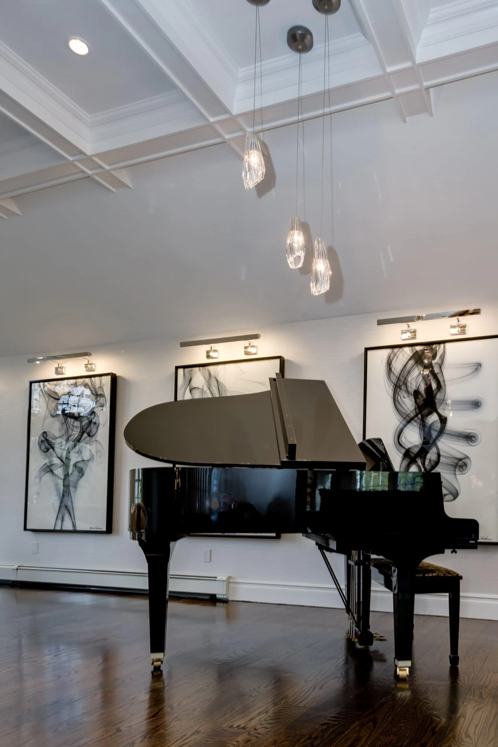 A grand piano sits on a polished wooden floor in a modern room with three abstract art pieces on the wall and pendant lights hanging from the ceiling.