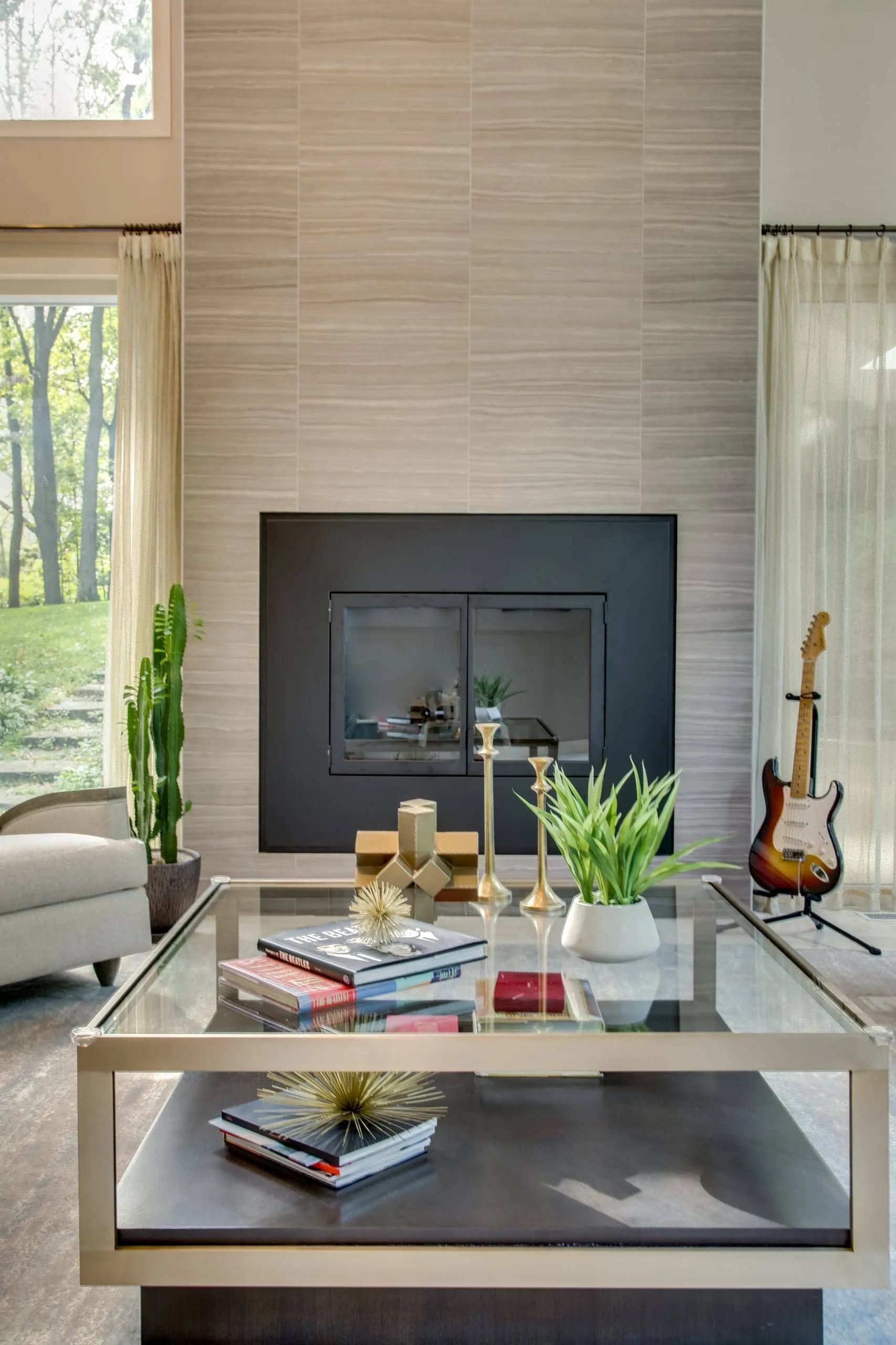 A modern living room with a glass coffee table holding books, gold decor, and potted plants, a black fireplace, tall windows with sheer curtains, a gray armchair, a cactus, and an electric guitar in the corner.