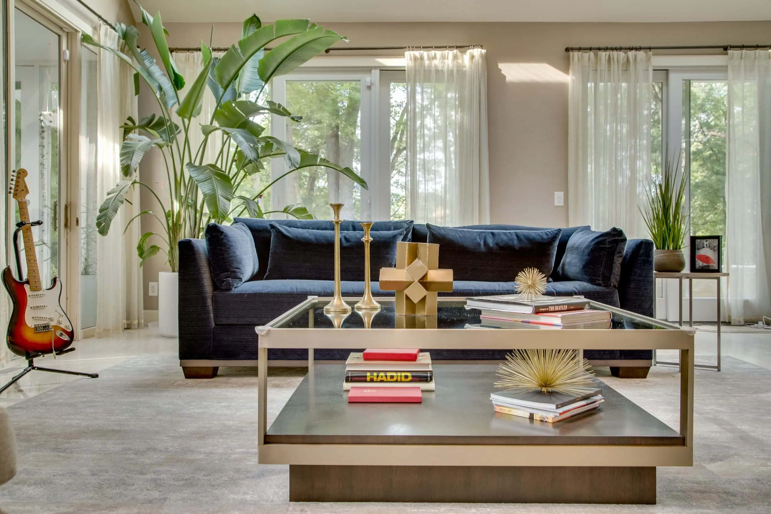 Modern living room with a dark blue sofa, large green plants, glass coffee table with books and gold decor, sheer curtains, and a red electric guitar in the corner near glass doors. Natural light fills the space.