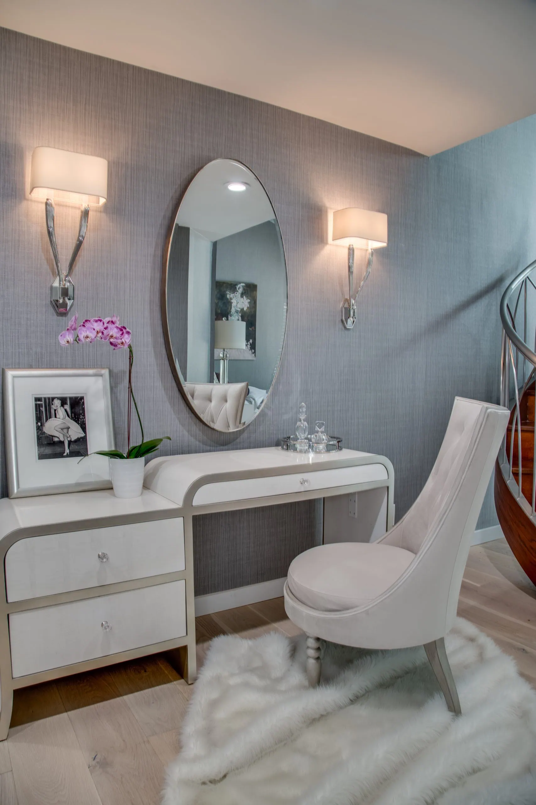 Elegant vanity area with a white desk, oval mirror, and cushioned chair on a fluffy rug. Decor includes a potted orchid, framed photo, and crystal bottles, with two modern wall sconces above.