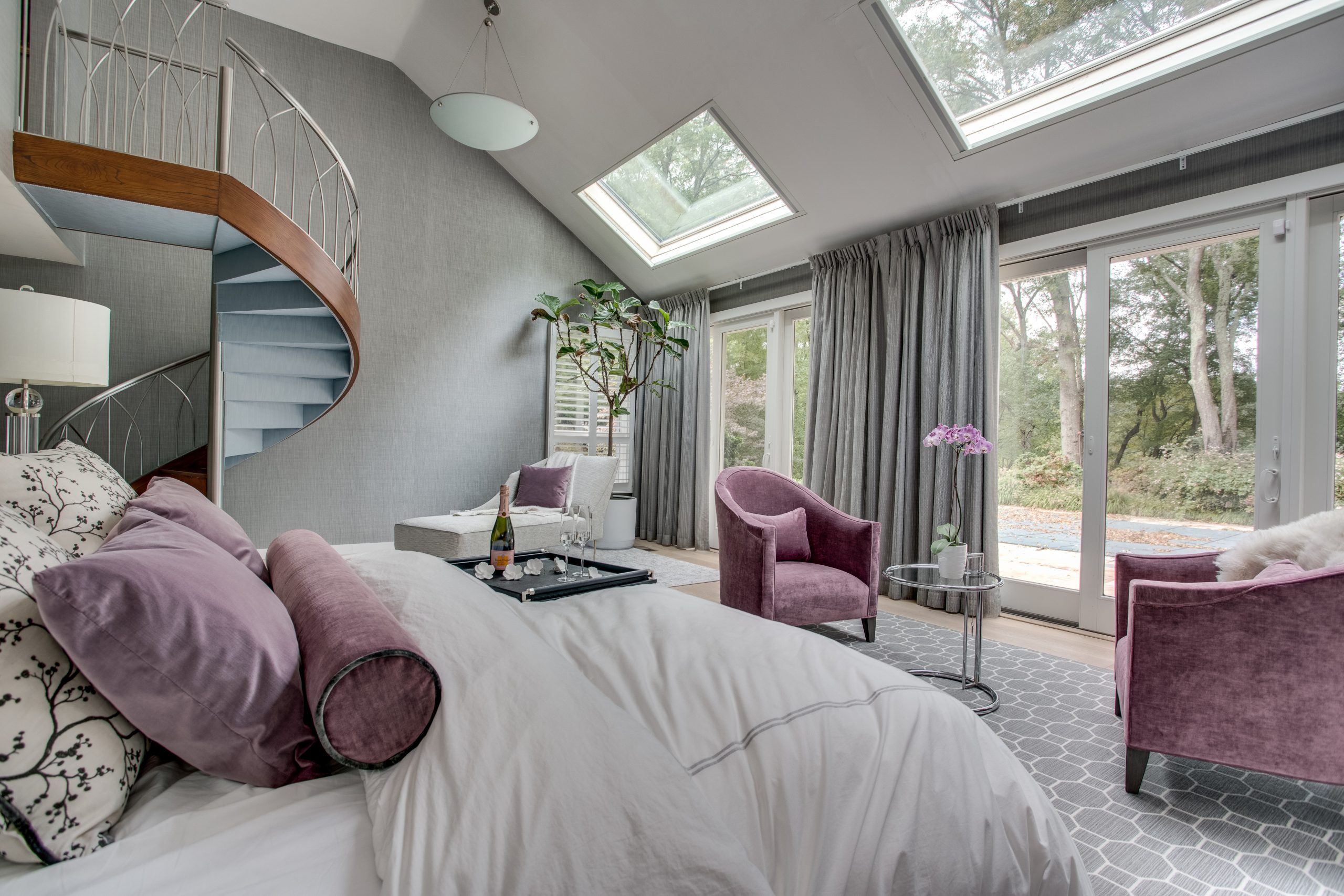 Modern bedroom with a spiral staircase, large skylights, and glass doors. The room features a bed with white and lavender bedding, purple chairs, a small table, and a potted plant. Lush trees are visible outside.