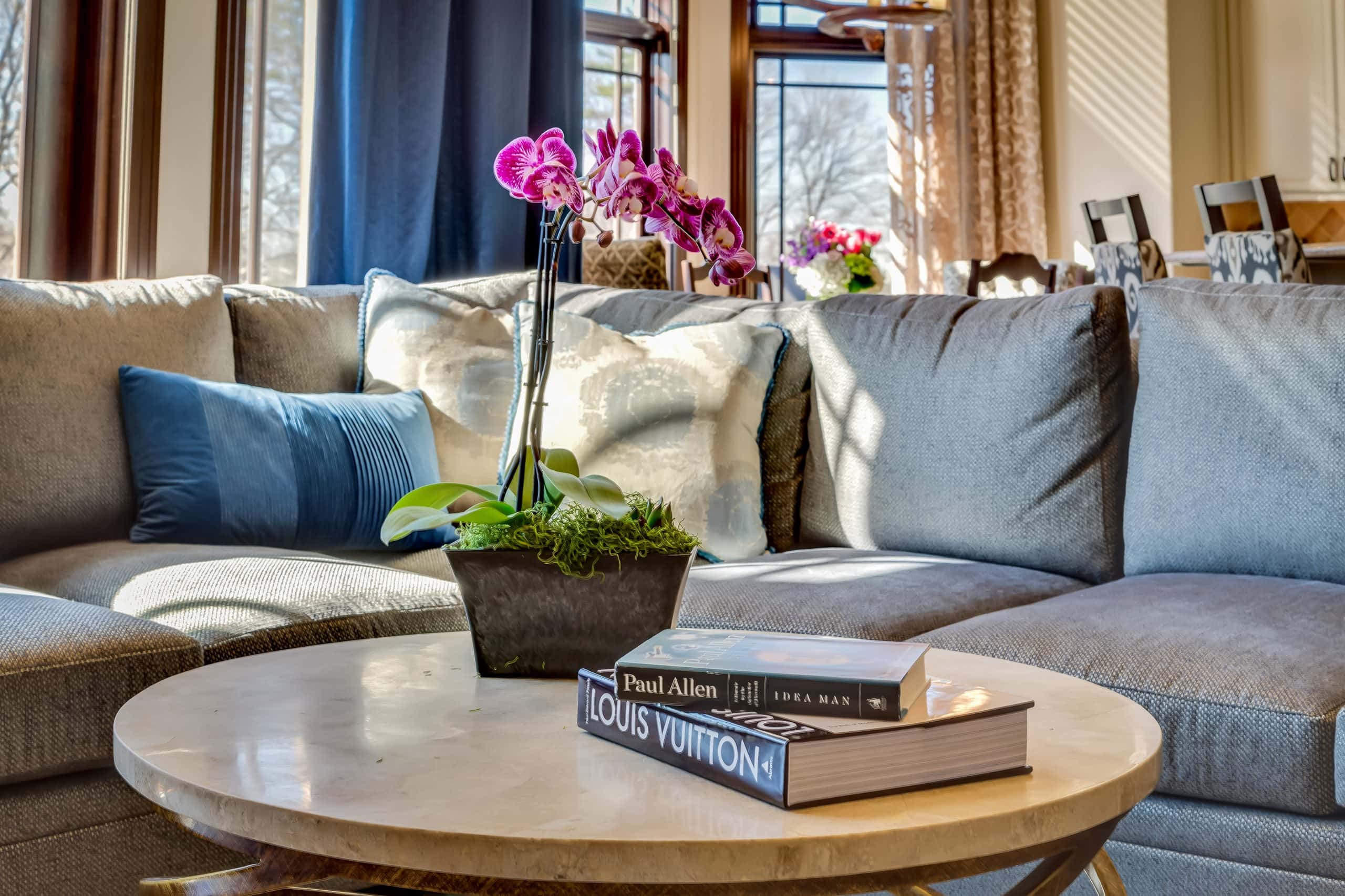 A cozy living room with a gray sectional sofa, decorative blue and white pillows, a potted orchid on a round marble coffee table, and two books stacked on the table near a window with natural sunlight.
