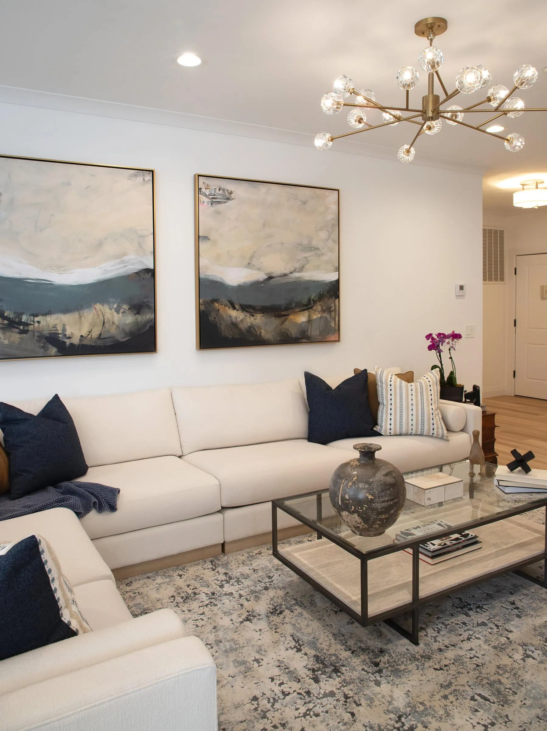 Modern living room with a white sectional sofa, blue and white cushions, abstract artwork on the wall, a glass coffee table with decor items, a patterned rug, and a contemporary chandelier overhead.