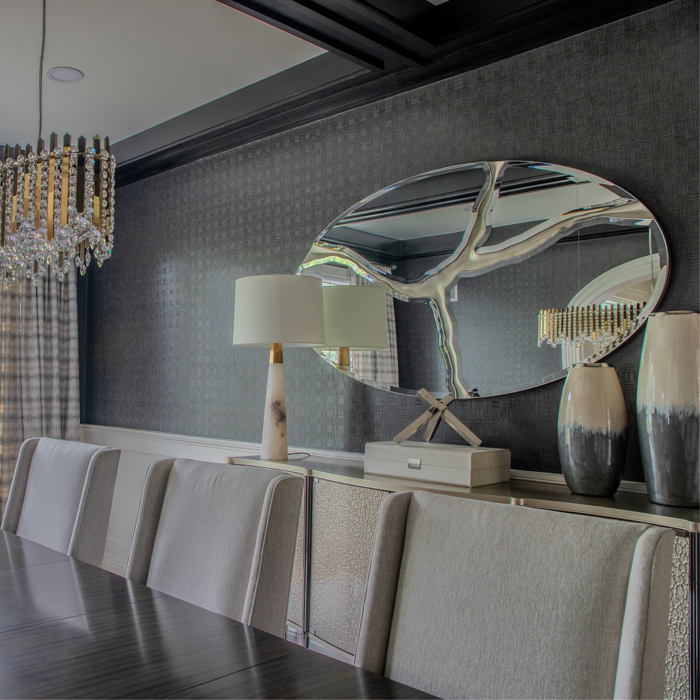 A modern dining room with light gray upholstered chairs, a dark wooden table, a sideboard with vases, a white lamp, and an oval mirror. A crystal chandelier and patterned curtains are reflected in the mirror.