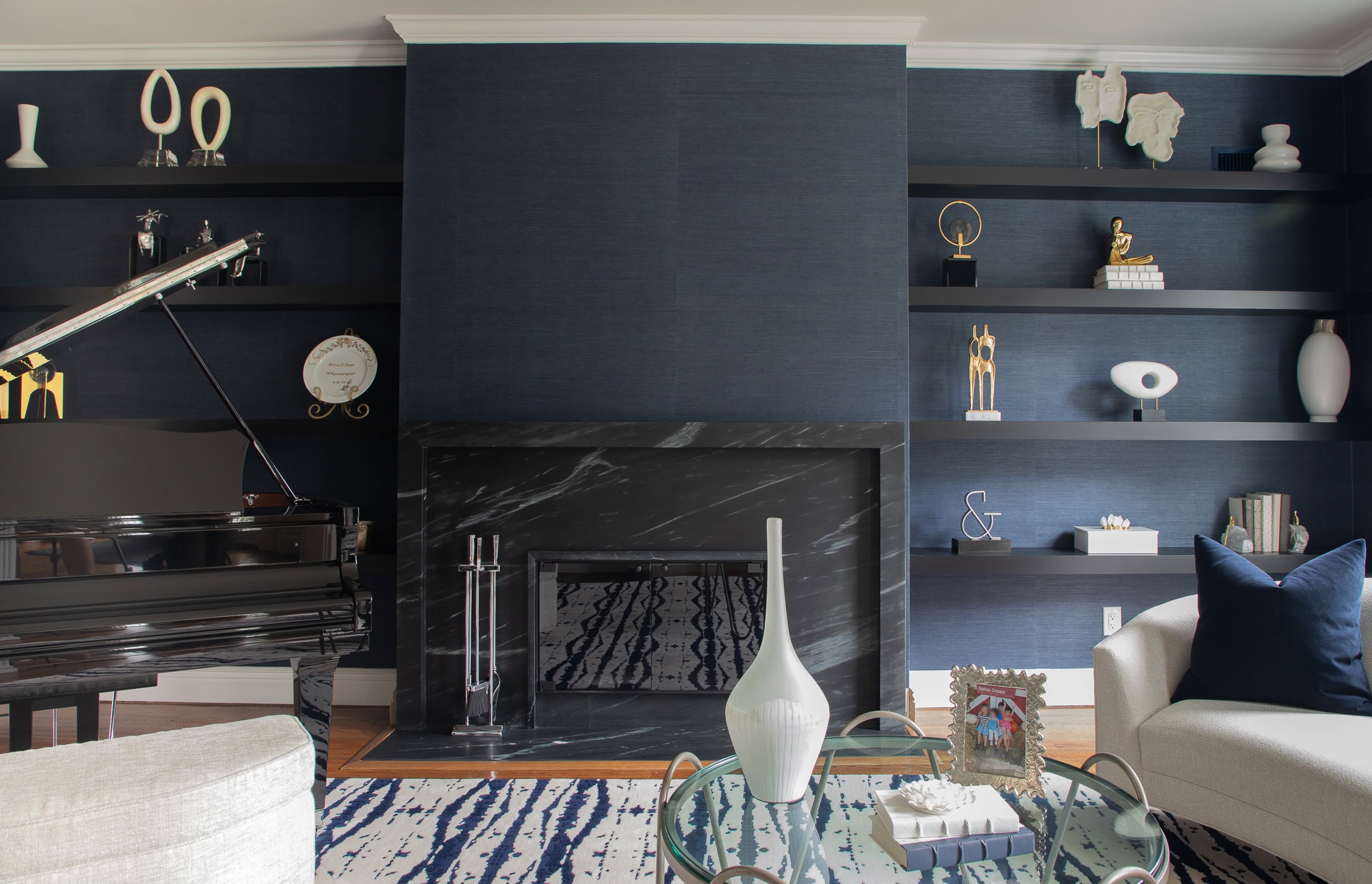 A modern living room with dark blue walls, black shelves holding decorative objects, a black fireplace, a grand piano, and contemporary furniture in neutral tones. A glass coffee table sits on a patterned rug.